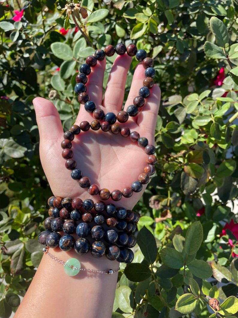 May include: A stack of six bracelets made of dark blue and brown tiger's eye beads. The bracelets are layered on a person's wrist, with a silver chain bracelet with a green jade charm on top.