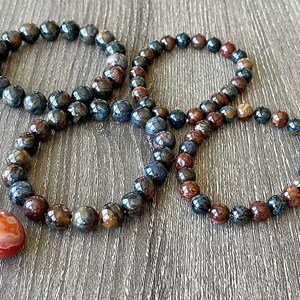 May include: Four bracelets made of blue and brown tiger's eye beads. The bracelets are stacked on top of each other on a wooden surface.