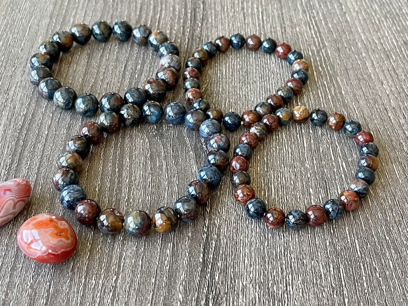May include: Four bracelets made of blue and brown tiger's eye beads. The bracelets are stacked on top of each other on a wooden surface.