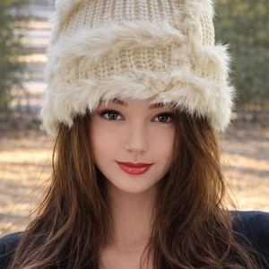 May include: A cream-coloured knitted hat with a faux fur trim. The hat is worn by a person with long brown hair and red lipstick. The background is blurred, with a natural outdoor setting.