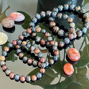 May include: Three beaded bracelets made of blue, brown, and gold stones. The bracelets are stacked on top of each other on a reflective surface. There are also four small, smooth stones in various shades of pink and orange.