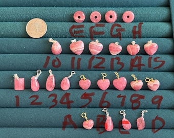 May include: A collection of 15 pink rhodochrosite gemstone pendants with silver and gold accents. The pendants are arranged in rows on a green surface with numbers and letters labeling each one.