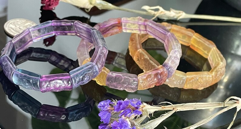 May include: Three colourful bracelets made of faceted glass beads. The beads are carved with intricate designs and feature a gradient of purple, pink, and yellow.