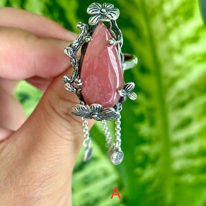 May include: A silver ring with a pink gemstone and floral details. The ring has dangling charms with floral designs.