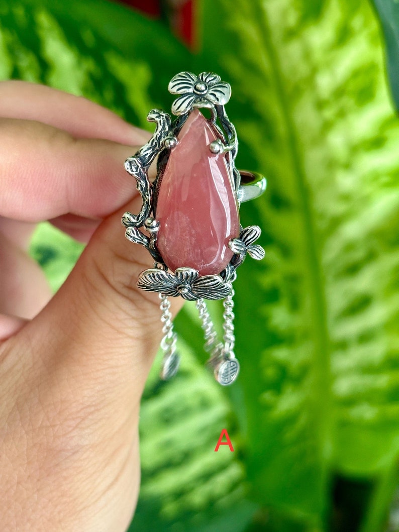 May include: A silver ring with a pink gemstone and floral details. The ring has dangling charms with floral designs.