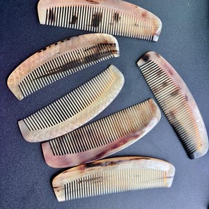 Puede incluir: Una colección de seis peines curvos para el cabello hechos de material de cuerno pulido. Cada peine presenta un patrón natural veteado único en tonos crema, bronceado y marrón oscuro. Algunos peines muestran una textura grabada en rombos.