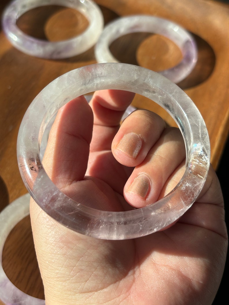 May include: A clear, translucent amethyst bangle bracelet with a light purple hue. The bracelet is made of natural stone and has a smooth, polished finish.