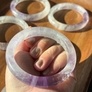 May include: Four amethyst bracelets with a light purple hue. The bracelets are stacked on top of each other, with the top bracelet being held by a hand.
