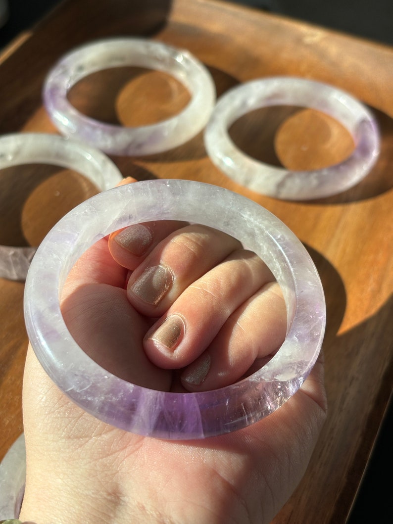 May include: Four amethyst bracelets with a light purple hue. The bracelets are stacked on top of each other, with the top bracelet being held by a hand.