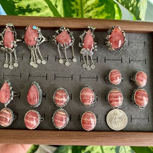 May include: A collection of 15 silver rings with pink and white striped gemstone settings. The rings are displayed in a wooden box with a gray felt lining.