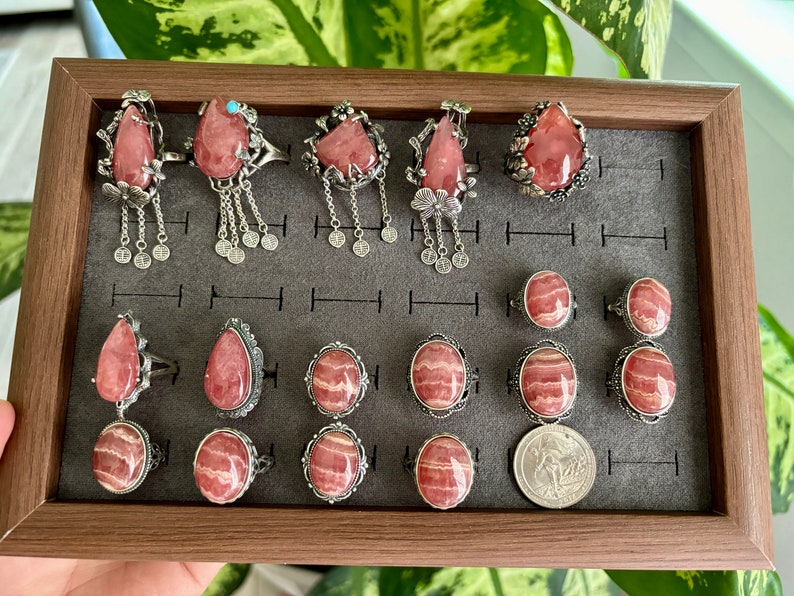 May include: A collection of 15 silver rings with pink and white striped gemstone settings. The rings are displayed in a wooden box with a gray felt lining.