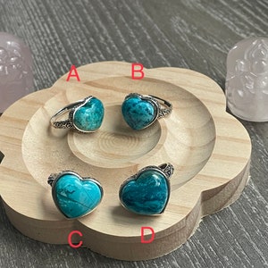 May include: Four silver rings with turquoise heart-shaped stones. The rings are displayed on a wooden surface with two pink carved stone decorations.