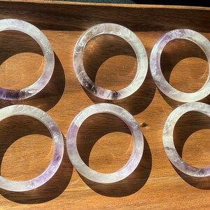 May include: Six clear amethyst crystal bracelets with a purple hue. The bracelets are stacked on a wooden surface.