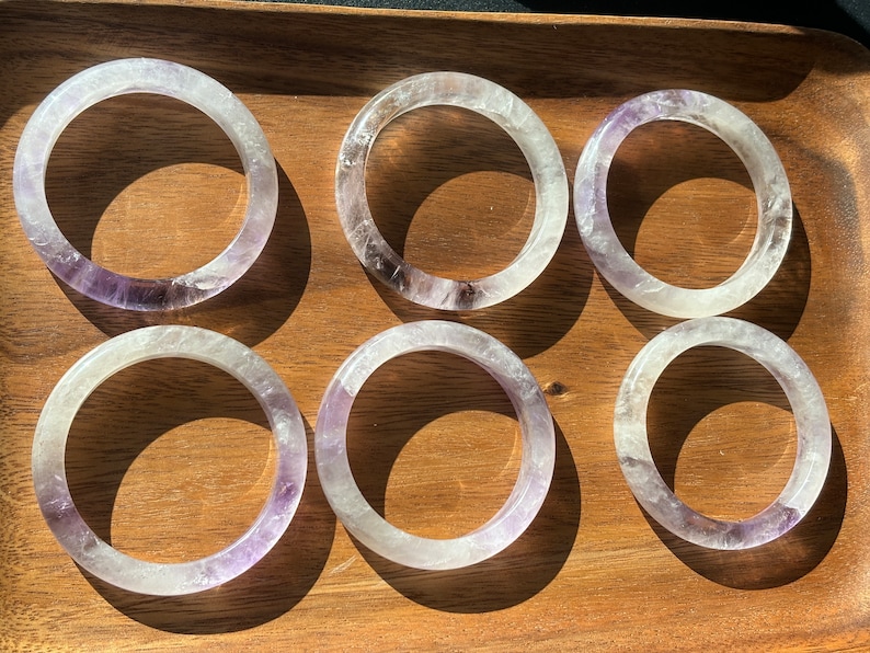 May include: Six clear amethyst crystal bracelets with a purple hue. The bracelets are stacked on a wooden surface.