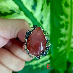 May include: A silver ring with a pink gemstone and floral accents. The ring is set against a green leafy background.