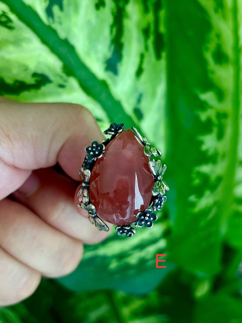 May include: A silver ring with a pink gemstone and floral accents. The ring is set against a green leafy background.