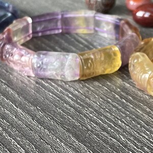 May include: Two bracelets made of carved, multi-coloured gemstone beads. The bracelet on the left is purple and the bracelet on the right is yellow.