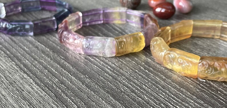 May include: Two bracelets made of carved, multi-coloured gemstone beads. The bracelet on the left is purple and the bracelet on the right is yellow.