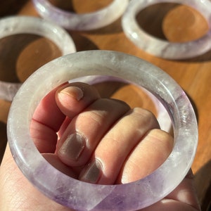 May include: A purple amethyst bangle bracelet with a white and purple color gradient. The bracelet is made of natural stone and has a smooth, polished finish.