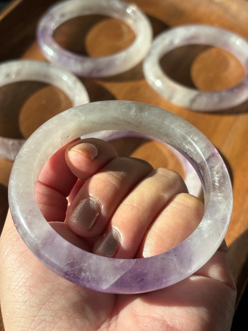 May include: A purple amethyst bangle bracelet with a white and purple color gradient. The bracelet is made of natural stone and has a smooth, polished finish.
