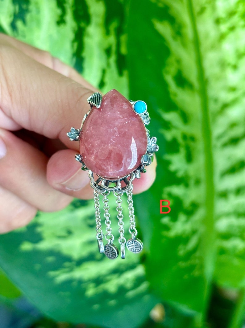 May include: A pink gemstone pendant with silver floral accents and a turquoise stone. The pendant has a chain with four charms.