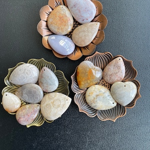 Puede incluir: Surtido de colgantes de piedra fosilizada en varias formas y tama&ntilde;os, incluyendo formas de l&aacute;grima, ovaladas y de coraz&oacute;n. Las piedras son una mezcla de marr&oacute;n claro, beige y blanco con marcas marrones y grises. Los colgantes se muestran en tres bandejas de metal decorativas.
