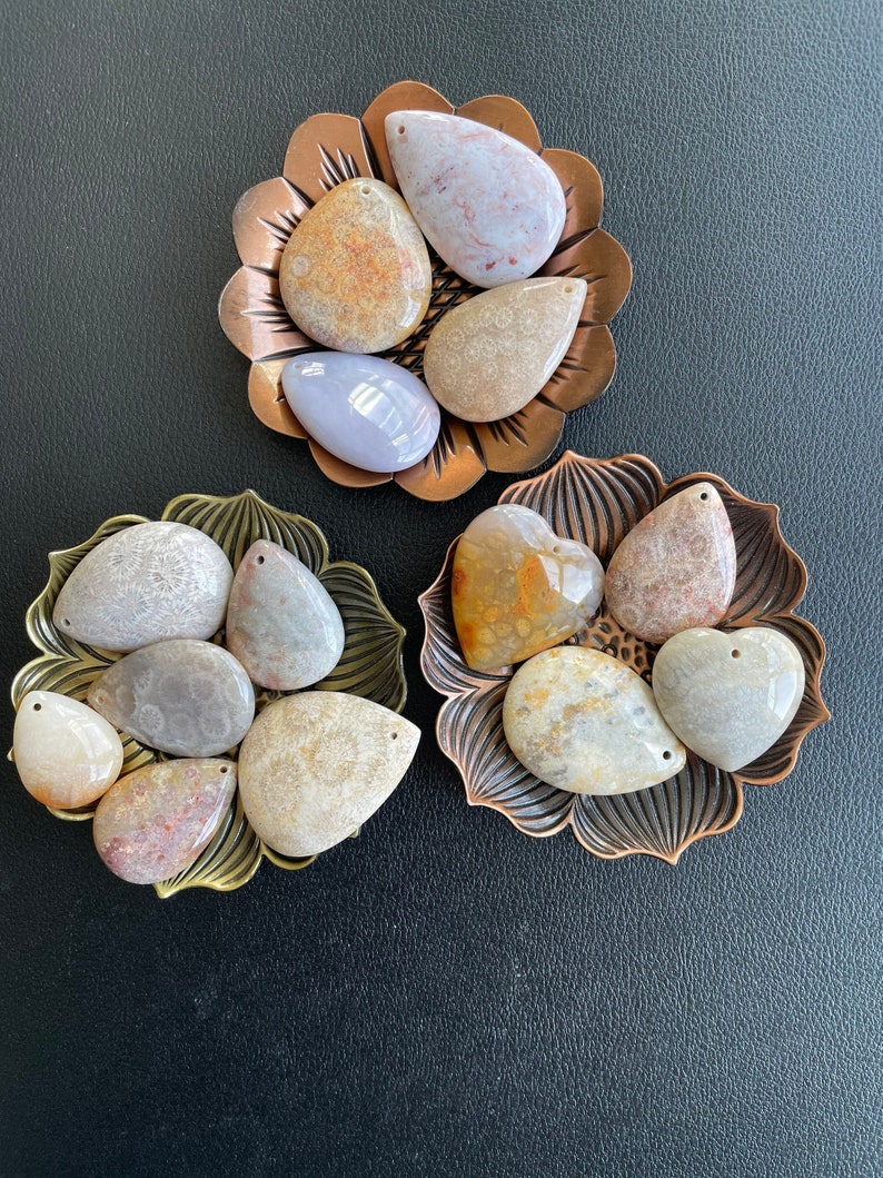 Puede incluir: Surtido de colgantes de piedra fosilizada en varias formas y tama&ntilde;os, incluyendo formas de l&aacute;grima, ovaladas y de coraz&oacute;n. Las piedras son una mezcla de marr&oacute;n claro, beige y blanco con marcas marrones y grises. Los colgantes se muestran en tres bandejas de metal decorativas.