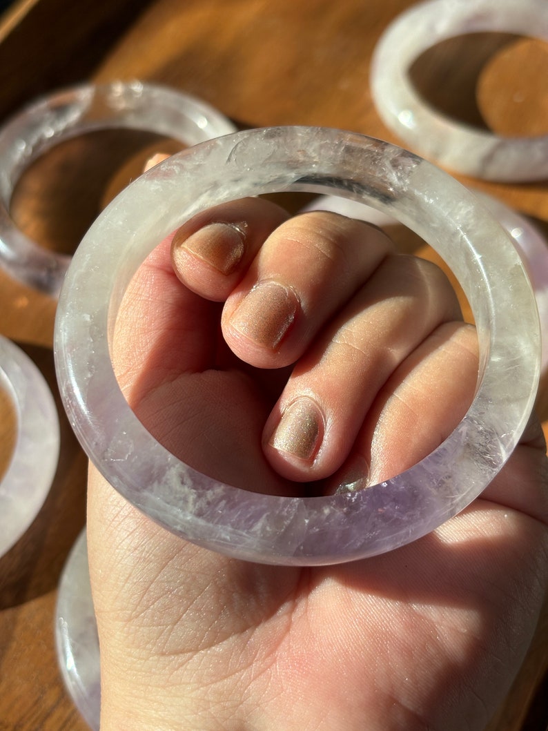 May include: A set of amethyst crystal bracelets. The bracelets are a light purple color and have a smooth, polished finish. They are stacked on top of each other, creating a beautiful display.