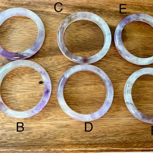 May include: Six amethyst bangle bracelets, labeled A through F, are displayed on a wooden tray. The bracelets are all made of clear amethyst with purple and white swirls.
