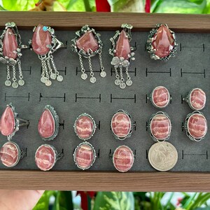May include: A collection of 15 silver rings with pink and white striped gemstone settings. The rings are displayed in a wooden box with a gray velvet lining.