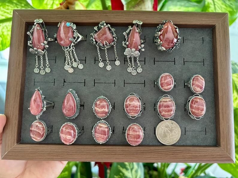May include: A collection of 15 silver rings with pink and white striped gemstone settings. The rings are displayed in a wooden box with a gray velvet lining.