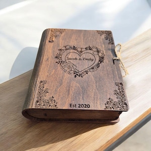 Könnte beinhalten: Eine holzfarbene, buchförmige Schatulle mit einem herzförmigen Blumendesign und den Namen "Jacob & Emily" auf der Vorderseite. Die Schatulle ist mit einer Schnur verschlossen und trägt die Gravur "Est 2020".