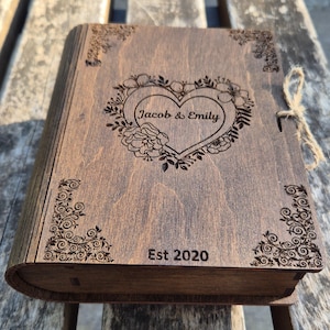 May include: A wooden keepsake box with a heart-shaped floral design and the names "Jacob & Emily" engraved on the front. The box is a warm brown color and has decorative corner accents. The year "Est 2020" is also engraved on the front.