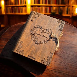 May include: A wooden box with a heart-shaped floral design and the names "Jacob & Emily" engraved on the front. The box is tied with twine and has the text "Est 2020" at the bottom. The box is on a wooden surface.