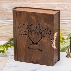 Puede incluir: Una caja de madera marrón oscuro con un diseño en forma de corazón tallado en la parte delantera. El diseño presenta dos árboles con ramas entrelazadas para formar un corazón. La caja está grabada con los nombres "Jacob & Emily" y la fecha "Est 2020".