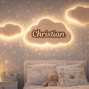 Könnte beinhalten: Kinderzimmer mit wolkenförmigen Wandleuchten und einem Bett. Die Leuchten sind beige mit warmem Schein, und die zentrale Wolke zeigt den Namen "Christian". Der Raum hat eine Tapete mit Sternenmuster, einen Teddybären und ein Hasen-Nachtlicht.