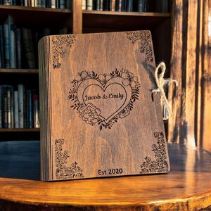 May include: A wooden book with a heart-shaped floral design and the names "Jacob & Emily" engraved on the cover. The book is tied with twine and has the text "Est 2020" at the bottom. The wood has a warm, natural tone.