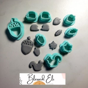 May include: A set of 14 teal plastic cookie cutters in the shape of acorns, leaves, and squirrels. The cutters are arranged on a white surface with gray clay cutouts.