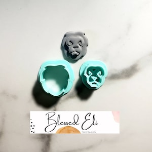 May include: Three polymer clay lion head cutters and a finished lion head. The lion head is gray, while the cutters are light blue. The cutters are in the shape of the lion's head. The image includes the text "Blessed Eli" on a banner.