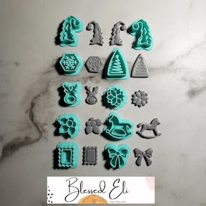以下が含まれることがあります： ターコイズとグレーのクリスマスをテーマにしたクッキー型18個セット。型には、木、雪片、トナカイ、馬、リボン、星など、さまざまな形が含まれています。型は白い表面に2列に並べて配置されています。画像の右下隅には「Blessed Eli」というテキストが表示されています。