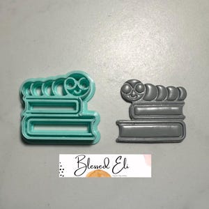 May include: Two cookie cutters: one teal, one grey. Each features a stack of books with a smiling caterpillar on top. The teal cutter is on the left, and the grey cutter is on the right. The cutters are for making biscuits.