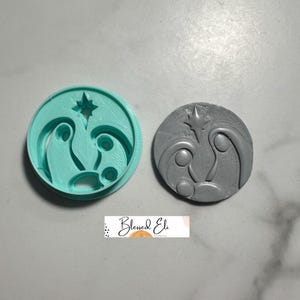 Puede incluir: Un cortador de galletas de plástico turquesa y una galleta gris. El cortador es redondo con una estrella y un diseño de escena de la natividad. La galleta es un relieve gris del mismo diseño. El texto "Blessed Eli" está en la parte inferior.