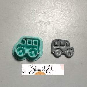 May include: Two school bus-shaped items. One is a teal-coloured cookie cutter, and the other is a grey, finished polymer clay charm. The cookie cutter has a detailed outline of a school bus with wheels and windows. The charm is a miniature version of the school bus.