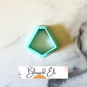 May include: A light blue plastic cookie cutter shaped like a diamond. The cutter is on a white marble surface. The text "Blessed Eli Polymer Clay Jewelry" is visible in the bottom right corner of the image.
