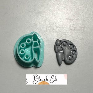 May include: A teal plastic cookie cutter shaped like a paint palette and brush, alongside a grey polymer clay paint palette and brush. The image includes the text "Blessed Eli".