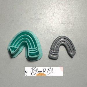 May include: Two rainbow-shaped polymer clay cutters. One is a light teal colour, and the other is grey. The cutters are on a grey surface. The text "Blessed Eli" is on a white banner.