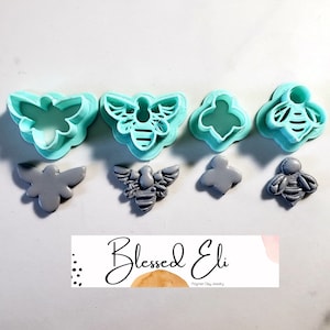 May include: Set of four bee-themed polymer clay cutters and four gray polymer clay bee shapes. The cutters are turquoise blue and the bee shapes are gray. The cutters are in the shape of a bee, a bee with wings, and a bee with a honeycomb pattern. The bee shapes are in the same shapes as the cutters. The image also includes a small business logo that says "Blessed Eli Polymer Clay Jewelry".