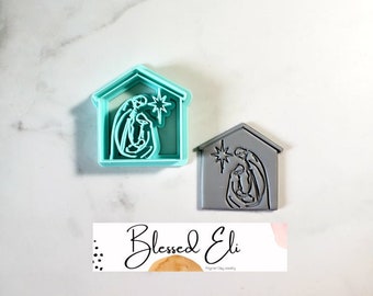 May include: Two cookie cutters in the shape of a house with a star and the Holy Family inside. The cutters are made of blue and gray plastic. The text "Blessed Eli" is visible on a white card below the cutters.