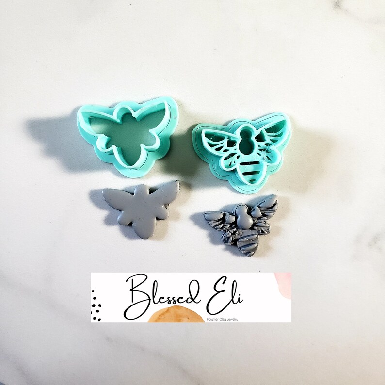 May include: Two turquoise plastic bee-shaped cookie cutters and two gray polymer clay bee shapes. The cookie cutters are detailed with wings, legs, and a striped abdomen. The clay bees are textured and have a three-dimensional appearance. The image also includes a small white label with the text "Blessed Eli Polymer Clay Jewelry".