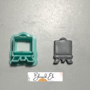 May include: Two cookie cutters shaped like an artist's easel. One is teal, the other is grey. The teal cutter has a square aperture. The grey cutter is a solid form. The text "Blessed Eli" is at the bottom.